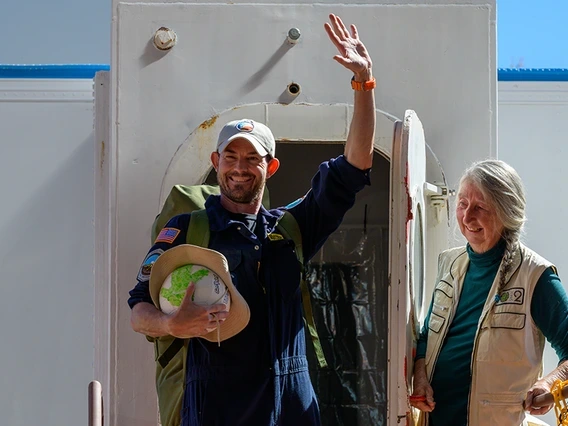 A person opens the door to a sealed habitat as a second person exits, waving and smiling.